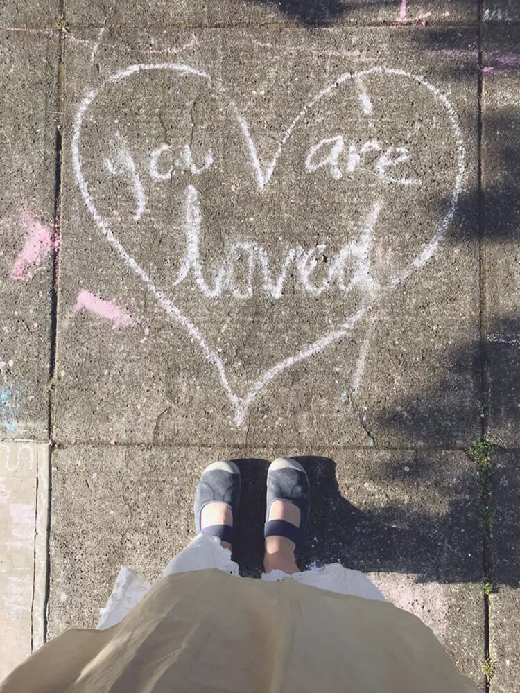 Kelly Rae Roberts standing over a chalk heart that reads 'you are loved'